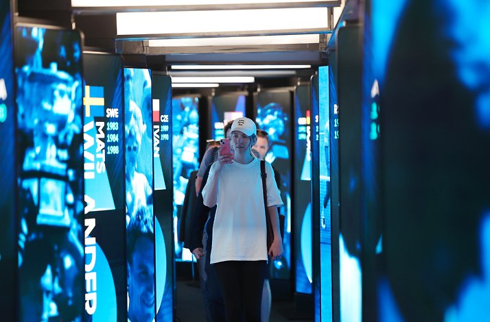 A visitor taking a video while walking down a hallway with glowing vertical screens of tennis player names.