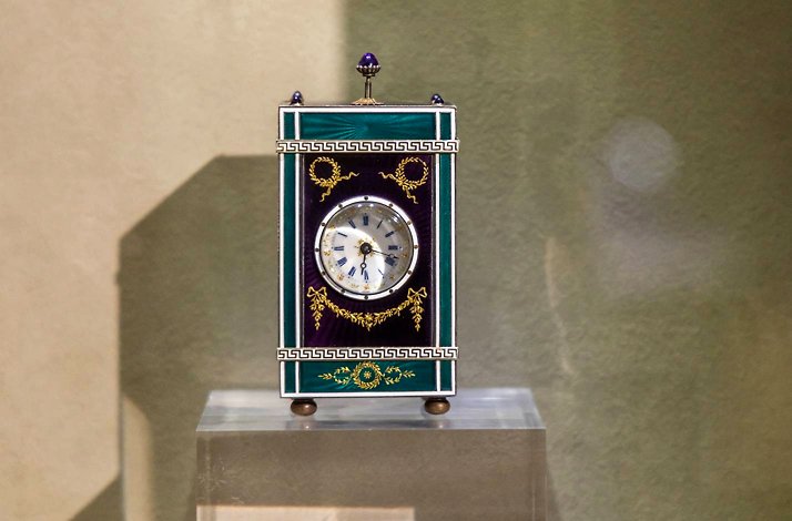 An ornate Cartier carriage clock with green and purple guilloché enamel and gold accents on a glass pedestal at Liang Yi Museum.
