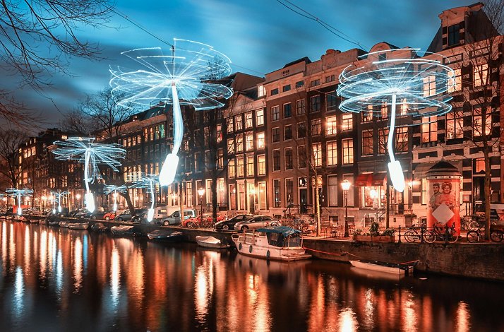 Illuminated dandelion-shaped light installations line a canal in a city with traditional buildings at dusk