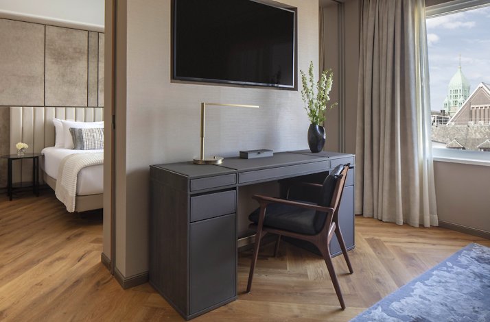 A hotel room with a desk, chair, and wall-mounted TV near a window.