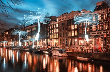 Illuminated dandelion-shaped light installations line a canal in a city with traditional buildings at dusk