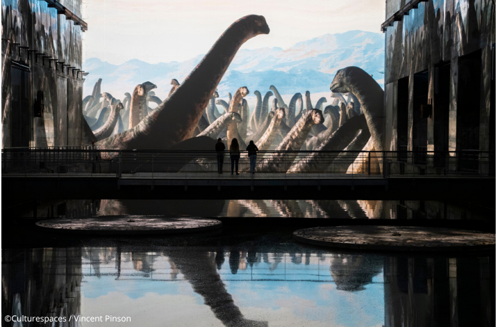 Visitors silhouetted on a bridge, surrounded by floor-to-ceiling projections of a sauropod herd at Bassins des Lumières. ©Culturespaces / Vincent Pinson