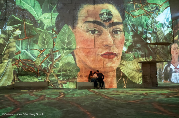 People sitting on a bench in a large, immersive digital art exhibition of the works of Frida Kahlo at Bassins des Lumières. ©Culturespaces / Geoffroy Groult
