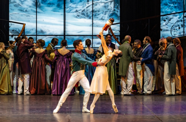 Ballet dancers perform a graceful lift onstage during a festive ballroom scene.