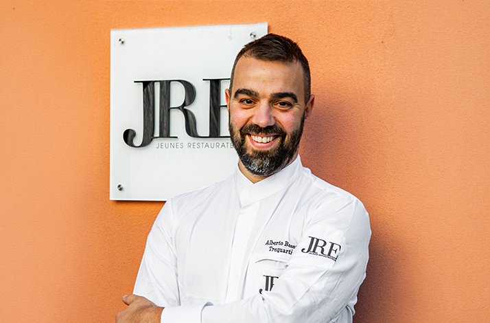 Chef Alberto Basso smiling in white JRE-branded chef's coat, arms crossed, posed before a 'JRE Jeunes Restaurateurs' wall plaque.