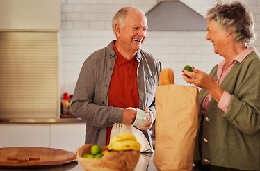 An old-couple smiling each other with a groceries bag in a table  