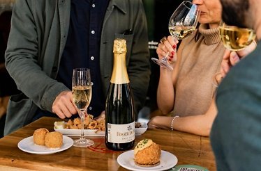 A man and a woman standing next to the table with bottle of wine with glasses and snacks on it.