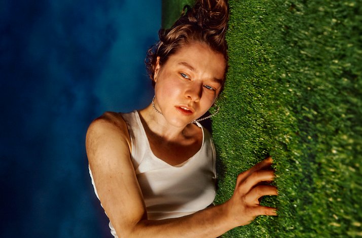 Emma D'Arcy in a white tank top, lying on green grass with a dramatic blue background for The Other Place promotion