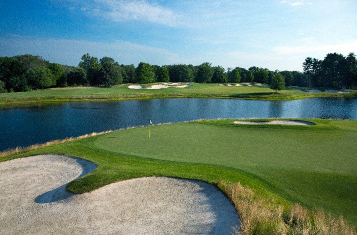 A golf course at TPC featuring a lake, surrounded by trees, under a clear blue sky.