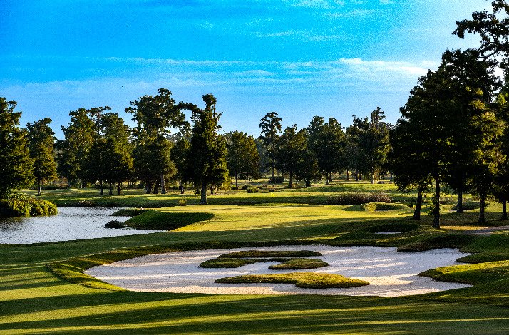 A golf course at TPC featuring a lake, surrounded by trees, under a clear blue sky.
