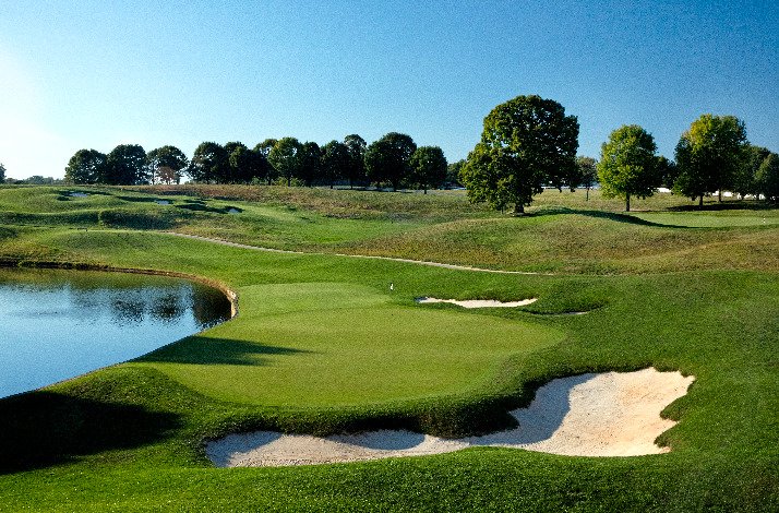 A golf course at TPC featuring a lake, surrounded by trees, under a clear blue sky.