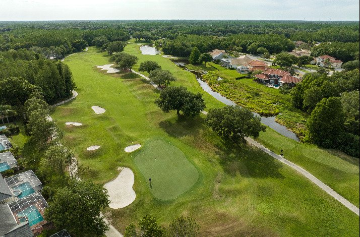 A bird's eye view of a TPC golf course, surrounded by trees and set under a clear blue sky.