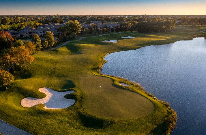 A bird's eye view of a TPC golf course, surrounded by trees and set under a clear blue sky.