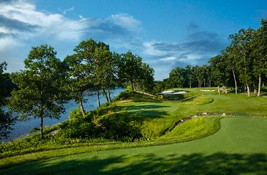 A golf course at TPC featuring a lake, surrounded by trees, under a clear blue sky.