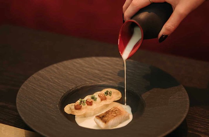 A refined dish being plated, with creamy sauce poured over a piece of seared fish and garnished crisps, presented on a dark plate in a fine dining setting.