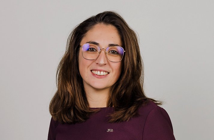 Stephanie Savarin with shoulder-length brown hair wearing a dark purple top with a small JRE logo, standing against a plain light background.
