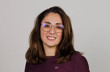 Stephanie Savarin with shoulder-length brown hair wearing a dark purple top with a small JRE logo, standing against a plain light background.