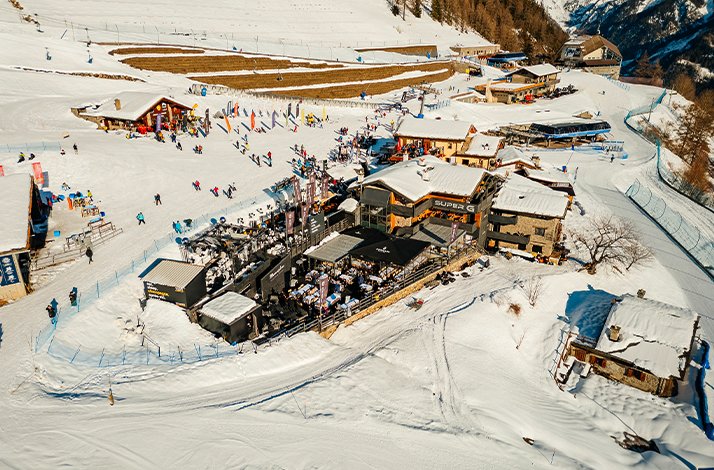 An aerial shot presents a ski resort village by the foot of Mont Blanc, featuring various buildings, slopes, and skiers.
