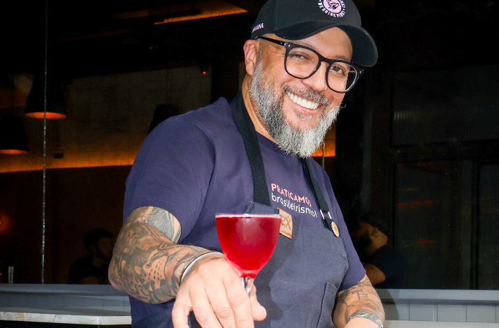 A bartender from Eximia holds a red cocktail in a stemmed glass inside a dimly lit bar or restaurant.