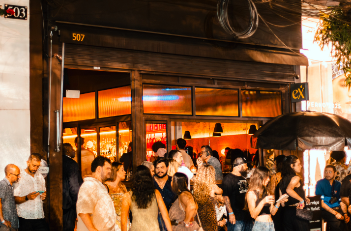 A lively crowd gathers outside a bar or restaurant, with warm lighting visible through large windows and people socializing near the entrance.