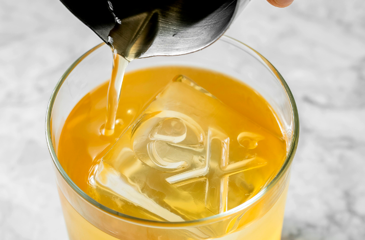 A close-up of a glass filled with an amber-colored drink, featuring a large ice cube engraved with the logo of the bar as liquid is being poured in.