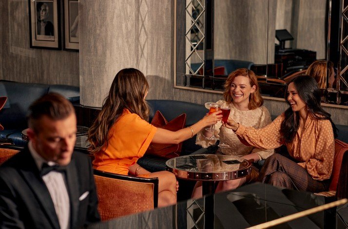 A group of womans sitting on a table enjoying cocktails with a musician playing piano wearing a smoking 