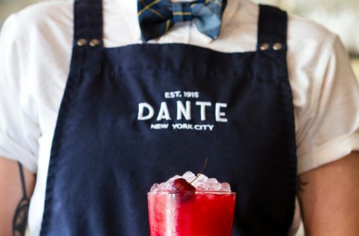 A bartender with a Caffe Dante apron presenting a red cocktail with a cherry