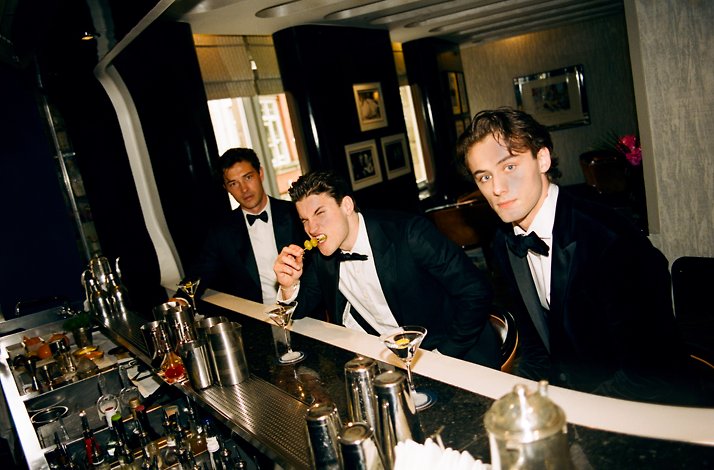 Three persons sitting next to a barman place with cocktails, one of them eating an olive of a Martini cocktail