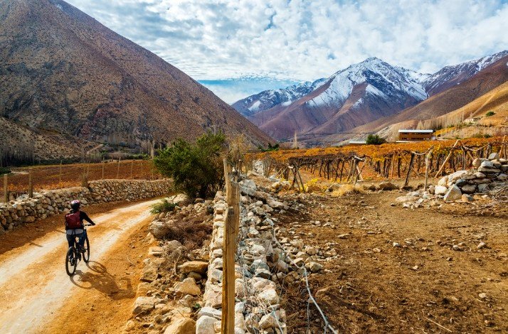  A man cycling along a dirt road surrounded by majestic mountains under a clear blue sky.