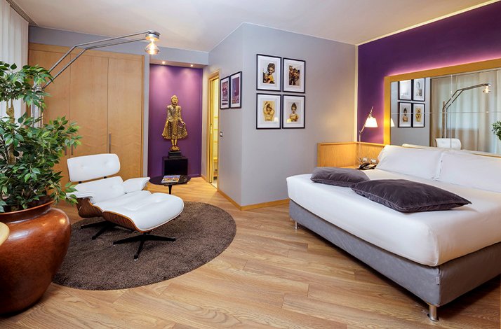 A luxurious hotel room for two with a large bed, a lounge chair and a purple, white and gold theme