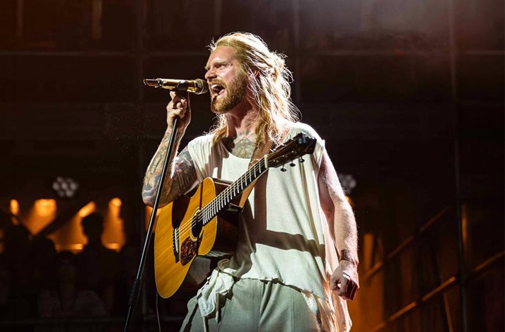 Tattooed performer singing passionately while playing acoustic guitar in Jesus Christ Superstar at an LW Theatre.