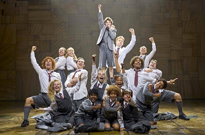 Ensemble cast of uniformed schoolchildren cheering and posing energetically on stage in Matilda The Musical.