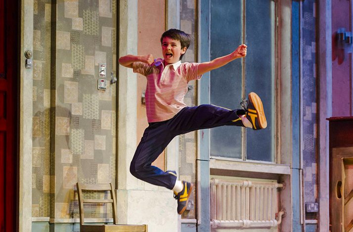 Young boy leaping mid-air with arms outstretched in an exuberant dance scene from Billy Elliot The Musical.