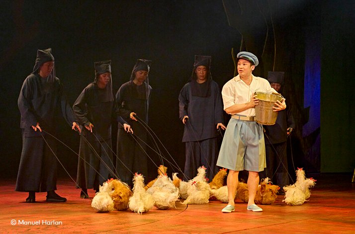 Performers in black operate chicken puppets as a boy holds a bucket on stage. ©  Manuel Harlan.