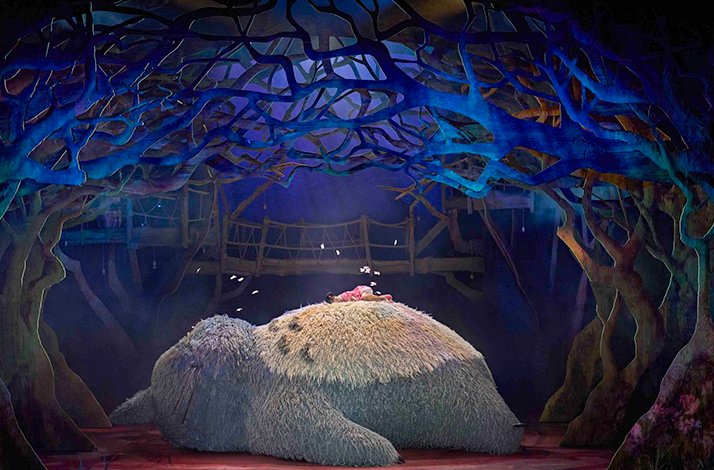 Totoro puppet lying down with a child resting on its belly under glowing trees.