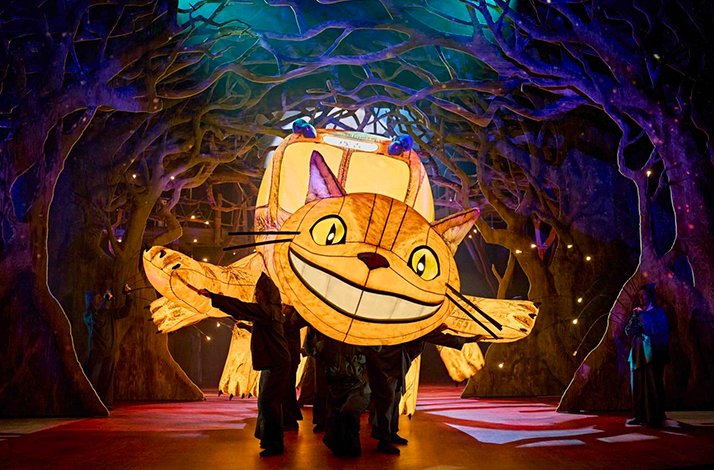Catbus puppet illuminated, operated by performers, in a magical forest scene.