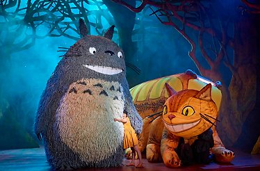 Totoro and Catbus puppets on stage with a performer in a forest setting.