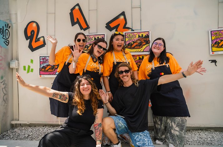 A group of six people smiling and posing together in front of colorful spray-paint artwork and graffiti-style numbers at an outdoor art workshop.
