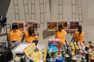 Four participants in yellow shirts creating colorful spray-paint artwork of race cars against a city skyline at an outdoor workshop, with spray cans and stencils on the table.