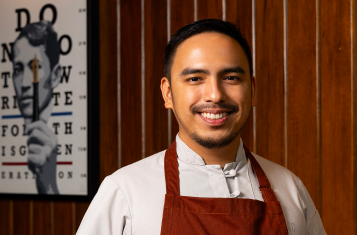 A smiling man wearing a chef's apron stands, exuding warmth and enthusiasm for cooking.