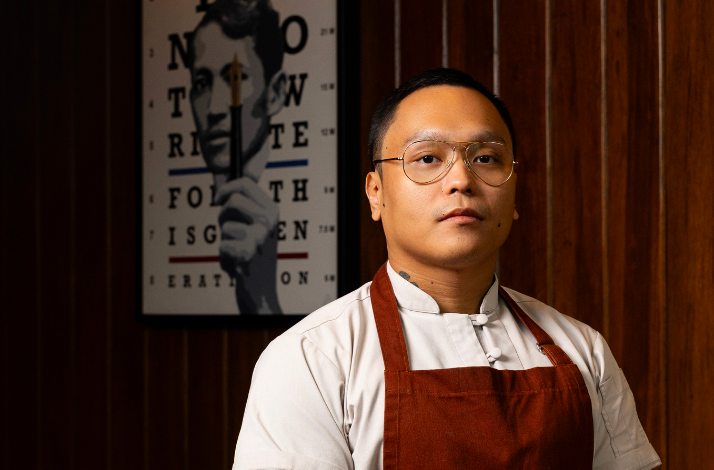 A man wearing an apron stands in front of a colorful painting, showcasing his artistic environment.