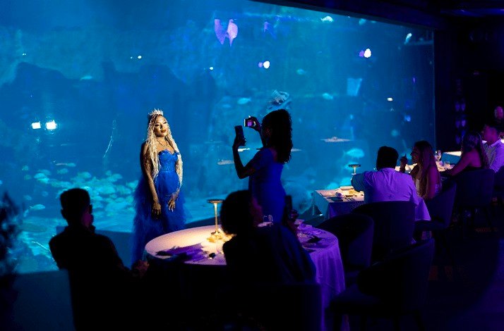A woman in a blue dress is being photographed in front of the aquarium at Su Va Na Restaurant.