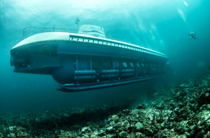 The submarine is fully submerged, cruising along the ocean floor with a diver nearby—highlighting the scale of the vessel