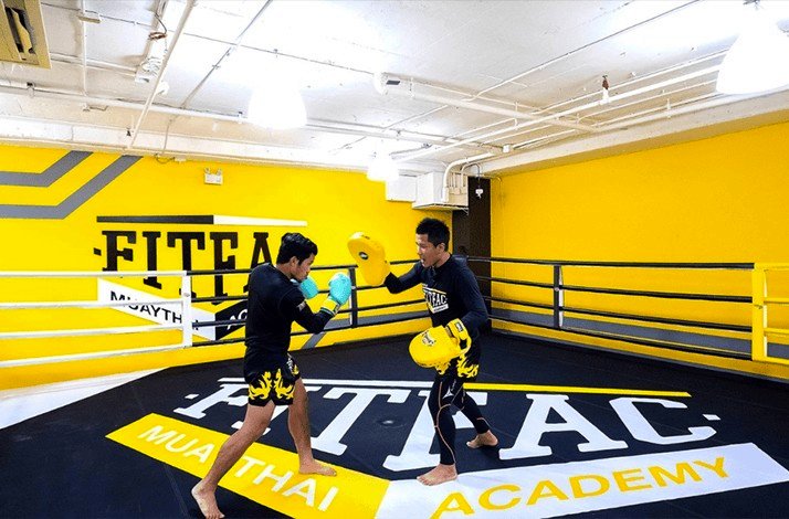 A Muay Thai student practices a kick with pads in a bright training gym.