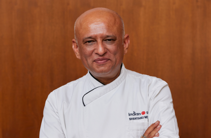 Chef Shantanu Mehrotra in white uniform standing against a wooden background.