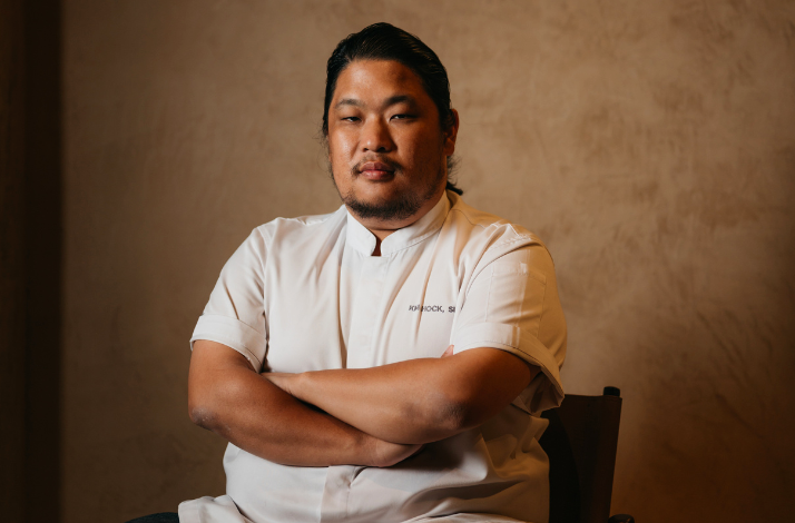 Chef Kim Hock Su in white uniform seated against a warm-toned wall.