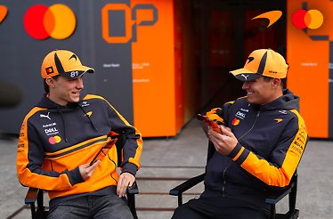 Lando Norris and Oscar Piastri are sitting and chatting wearing McLaren F1 team hoodies and caps.