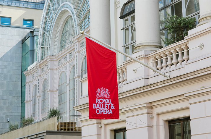 Daytime outdoor view of the Royal Ballet & Opera building showcasing a large red flag with the Royal Ballet & Opera logo.