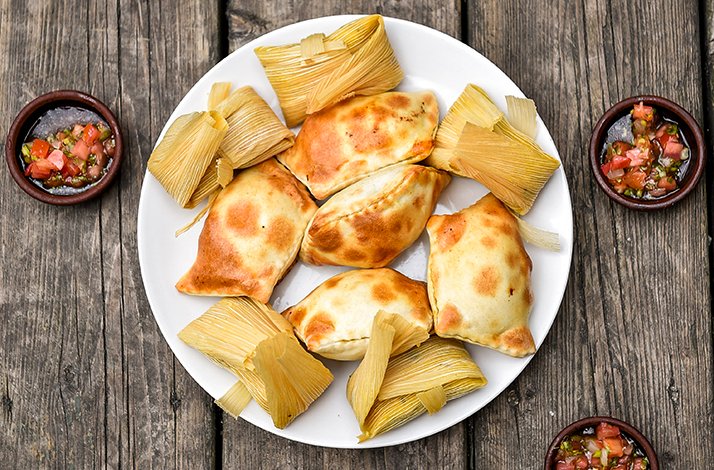 White plate holding golden-baked empanadas and corn husk-wrapped tamales, with small bowls of fresh salsa on a rustic wood surface.