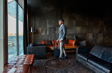 A man in a suit with a suitcase stands in the airport lounge area.